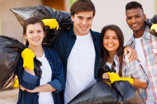 A group of four young adults standing outdoors in a residential area during daytime, each holding black rubbish bags filled with waste, positioned in front of a paved driveway or walkway. The individuals are smiling and appear to be participating in a community litter pick or rubbish removal activity. They are dressed casually, with some wearing yellow cleaning gloves, and are standing close together, indicating teamwork. In the background, there is a garden or front yard with blurred greenery and part of a building structure visible. The scene depicts a responsible approach to rubbish collection, suitable for local rubbish removal services such as those offered in Thamesmead, with the focus on environmental cleanliness and community engagement.