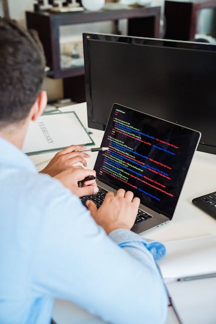 A close-up photograph of a laptop displaying lines of computer code on the screen, placed on a wooden desk surface. To the right of the laptop, there is a ceramic mug with a light brown finish, featur