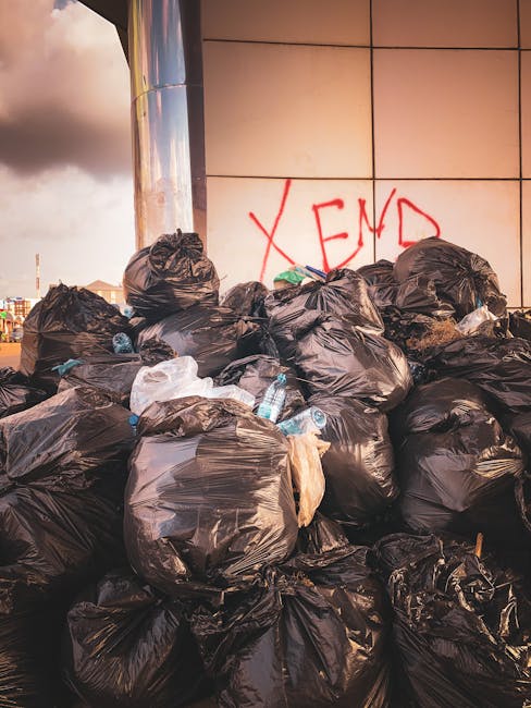 A large accumulation of black plastic rubbish bags filled with waste, piled against a modern tiled wall outdoors. Some of the bags are slightly bulging, indicating they are full, and a few are torn or