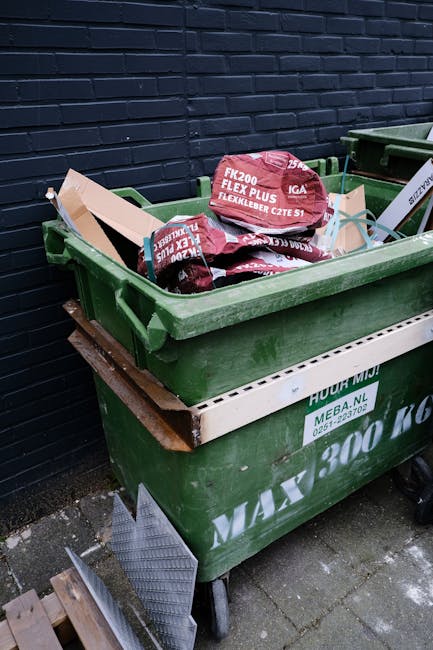 Two large black garbage bags filled with waste are positioned on the pavement in front of a wooden fence with vertical slats and a dense arrangement of leafy bushes or trees behind it. The bags appear
